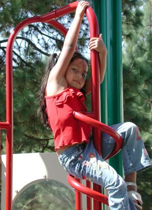 A little Hispanic girl holding onto red playground equipment, pine trees in background